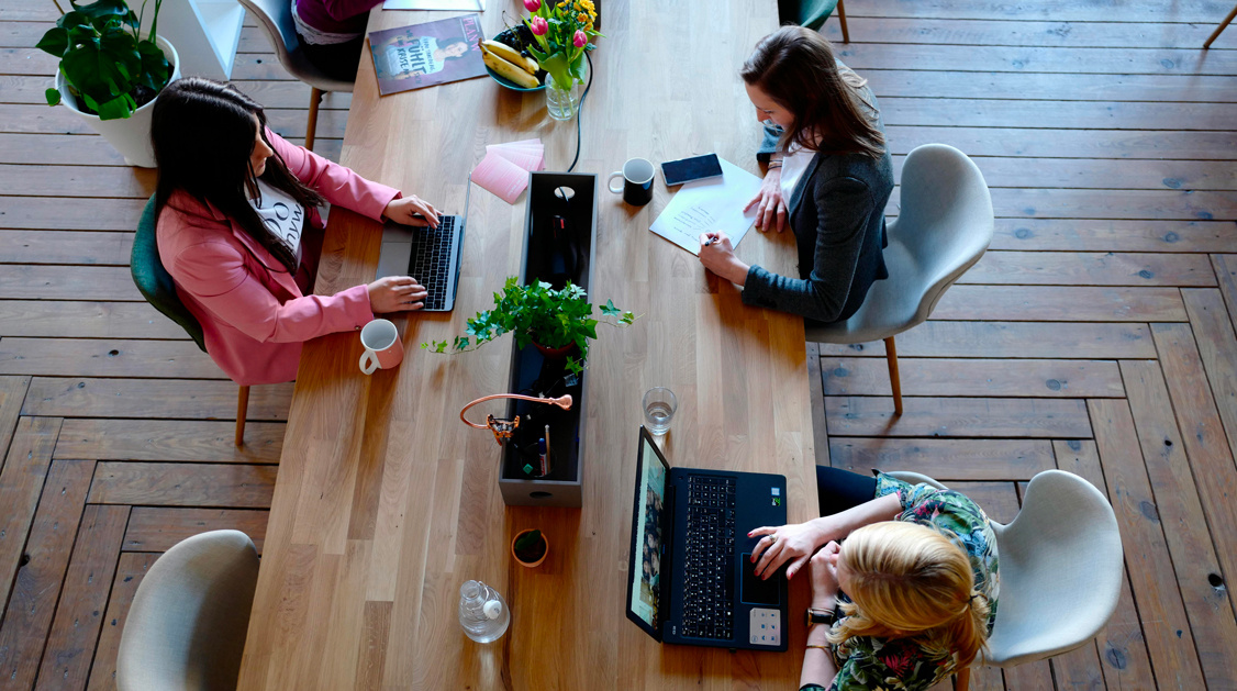 Vue de dessus de trois femmes travaillant sur des ordinateurs portables autour d'une grande table en bois dans un bureau.